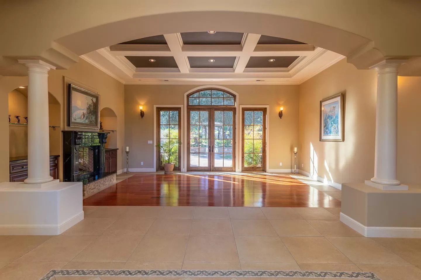 Spacious living room with large windows, coffered ceiling, and glass doors—perfect inspiration for your next Pensacola remodeling or home construction project. Natural sunlight highlights tiled floors, hardwood area, framed artwork, and elegant columns.