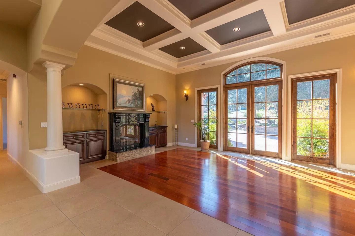 Spacious, sunlit living room with a coffered ceiling, large arched windows, hardwood floors, built-in cabinetry, and a fireplace—crafted by an expert general contractor. Light streams in through the windows, illuminating the elegant space.
