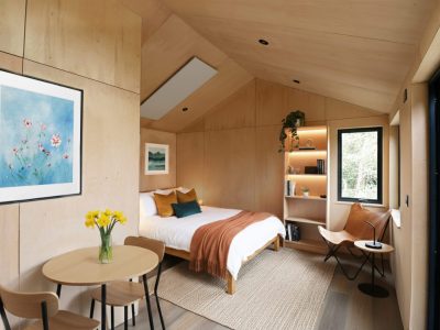 A cozy, modern bedroom with light wood walls, a double bed with orange and white bedding, a round table with yellow flowers, wall art, open shelving with plants and books, and large windows letting in natural light—perfect inspiration for your next Pensacola remodeling project.