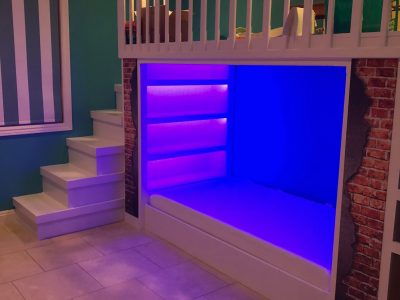 A modern, white bunk bed with stairs and built-in shelves is illuminated by blue LED lights. Perfect for a Pensacola remodeling project, the bed features a brick pattern on the sides, with teal walls and tiled flooring completing the look.
