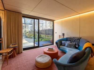 A cozy living room with a curved blue sofa, orange ottoman, pouf, and wooden chairs. Large sliding glass doors reveal a patio and garden outside. Light wood walls and a pink rug show the inviting style possible with Pensacola remodeling.