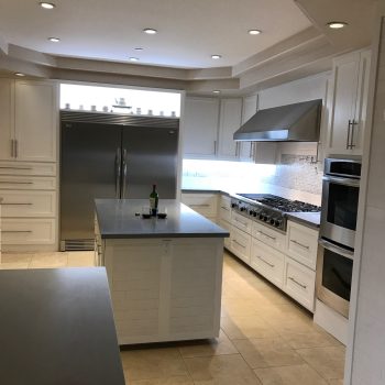 Modern kitchen with white cabinets, stainless steel appliances, a large fridge, double oven, gas range with vent hood, and central island. Expertly finished by a Pensacola remodeling general contractor. Beige tile flooring and recessed lighting.