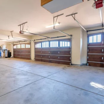 Spacious three-car garage with polished concrete floors, wooden garage doors with windows, ceiling-mounted openers, and wall-mounted shelving. Perfect for any home construction or remodeling project in Pensacola, this garage is well-lit with fluorescent lights.